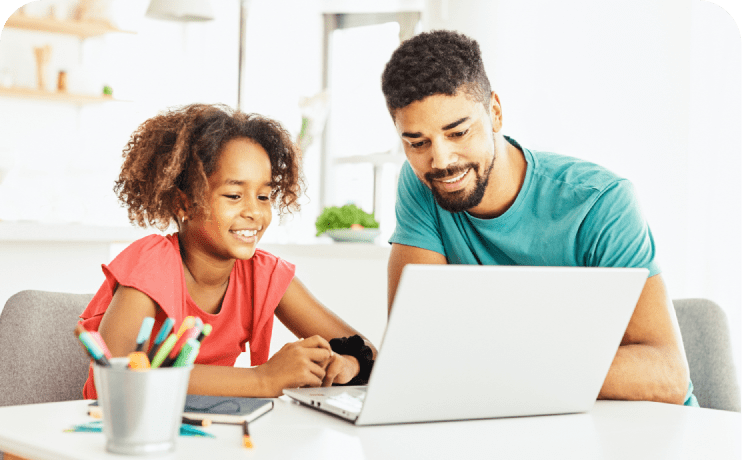 A man and a young girl are seated at a table, engaged with a laptop in front of them, sharing a moment of learning.