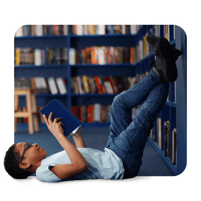 A boy reclines on the floor, gazing at a bookcase filled with various books and colorful decorations.